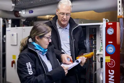 Zwei Techniker von Primeo Energie in Arbeitskleidung und mit Werkzeugen stehen vor einer Schalttafel in einer Industrieanlage. Eine Frau hält Dokumente, während der Mann auf etwas im Dokument zeigt.