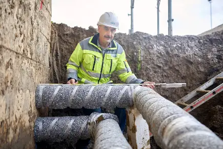 Primeo Energie Mitarbeiter in gelb-grauer Sicherheitskleidung und Schutzhelm bei der Arbeit an einer Wärmeleitung. Er steht in einem Erdgraben und hält ein Messgerät, während er die grossen isolierten Rohre prüft.