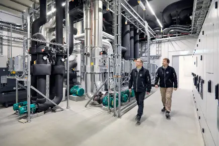 Zwei Techniker von Primeo Energie gehen durch die industrielle Heizzentrale von BaseLink mit grossen Rohrleitungen, Ventilen und Maschinen und prüfen die technischen Einrichtungen.