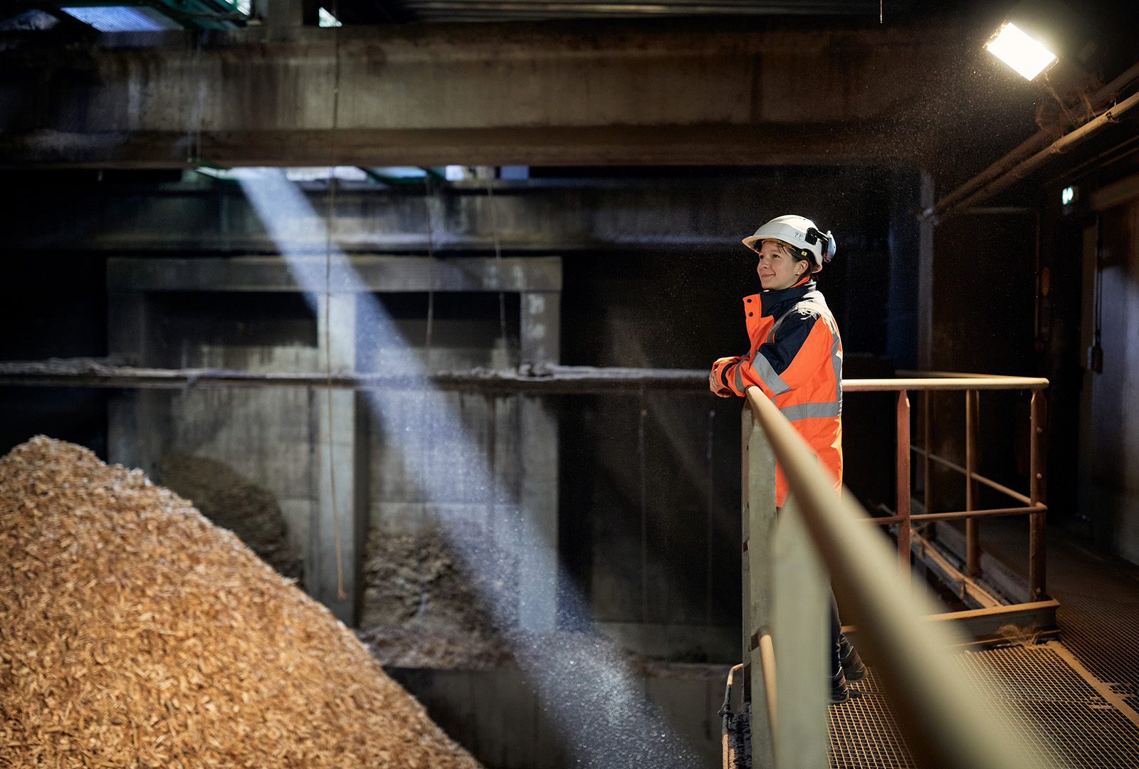 A Primeo employee in work clothes and hard hat stands on a raised metal aisle of a woodchip plant and looks at a large pile of woodchips.