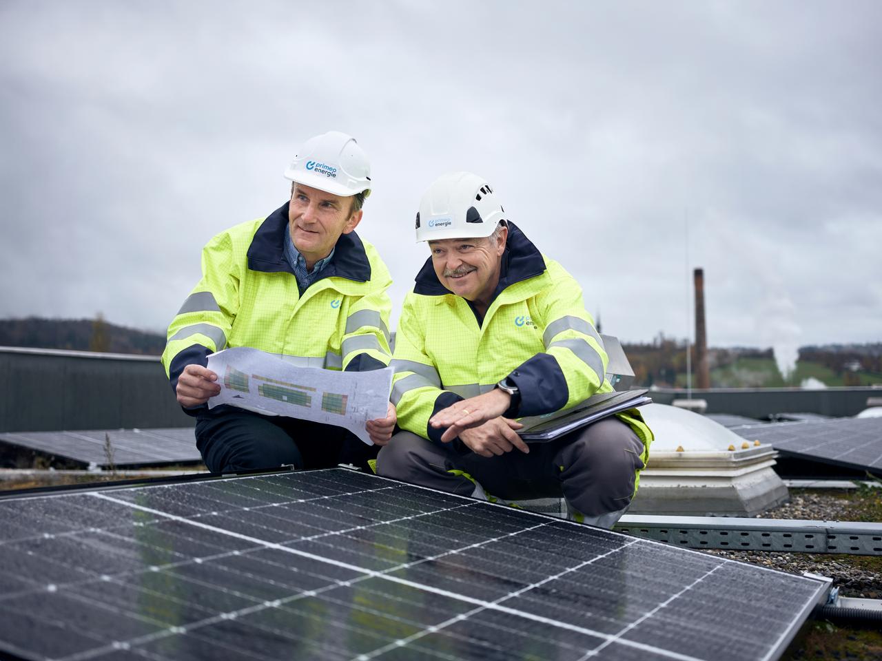  Two Primeo Energie engineers in high-visibility waistcoats and helmets inspect a solar installation on a roof. They study construction plans and note down details. 
