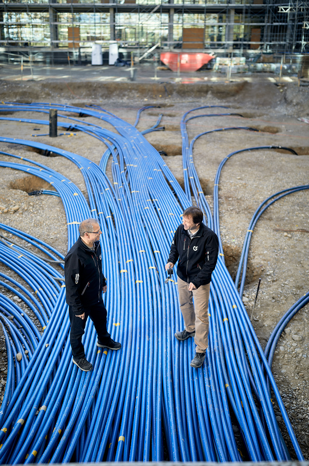 Two Primeo Energie technicians stand on the BaseLink construction site, surrounded by numerous blue underground pipes running along the ground. The building under construction with scaffolding can be seen in the background. 