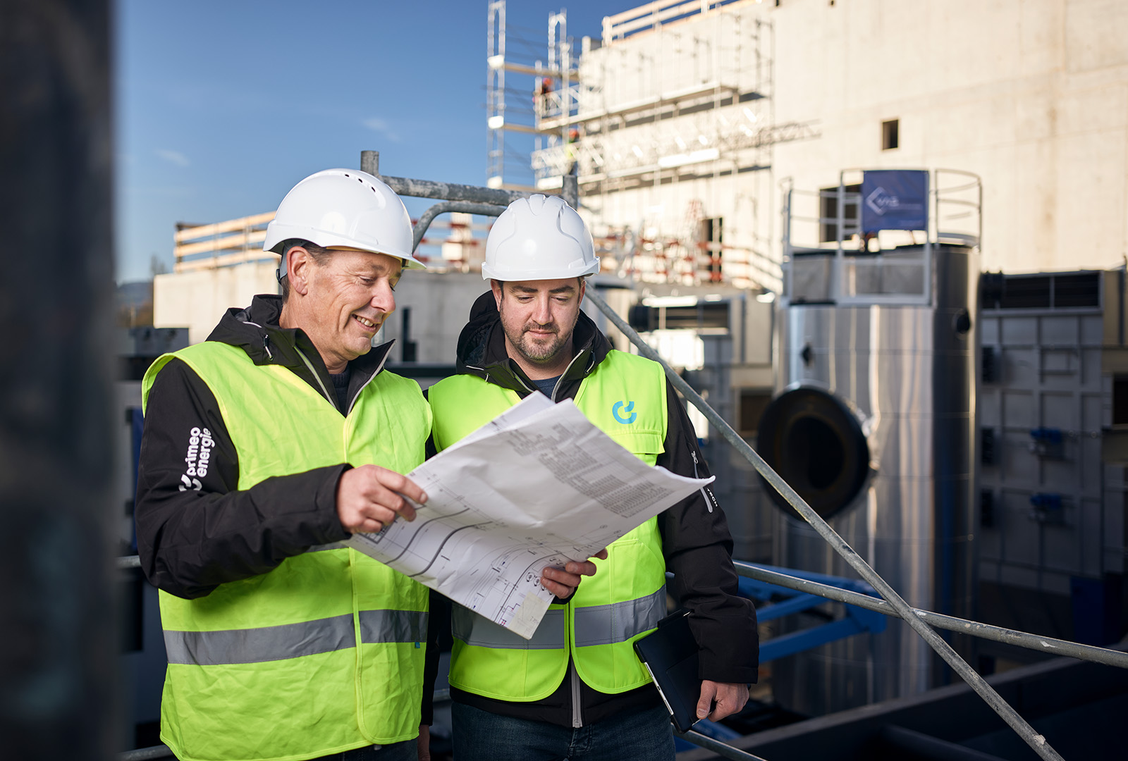 Two construction workers from Primeo Energie wearing hard hats and high-visibility waistcoats discuss construction plans on a building site. Scaffolding and industrial plants can be seen in the background.